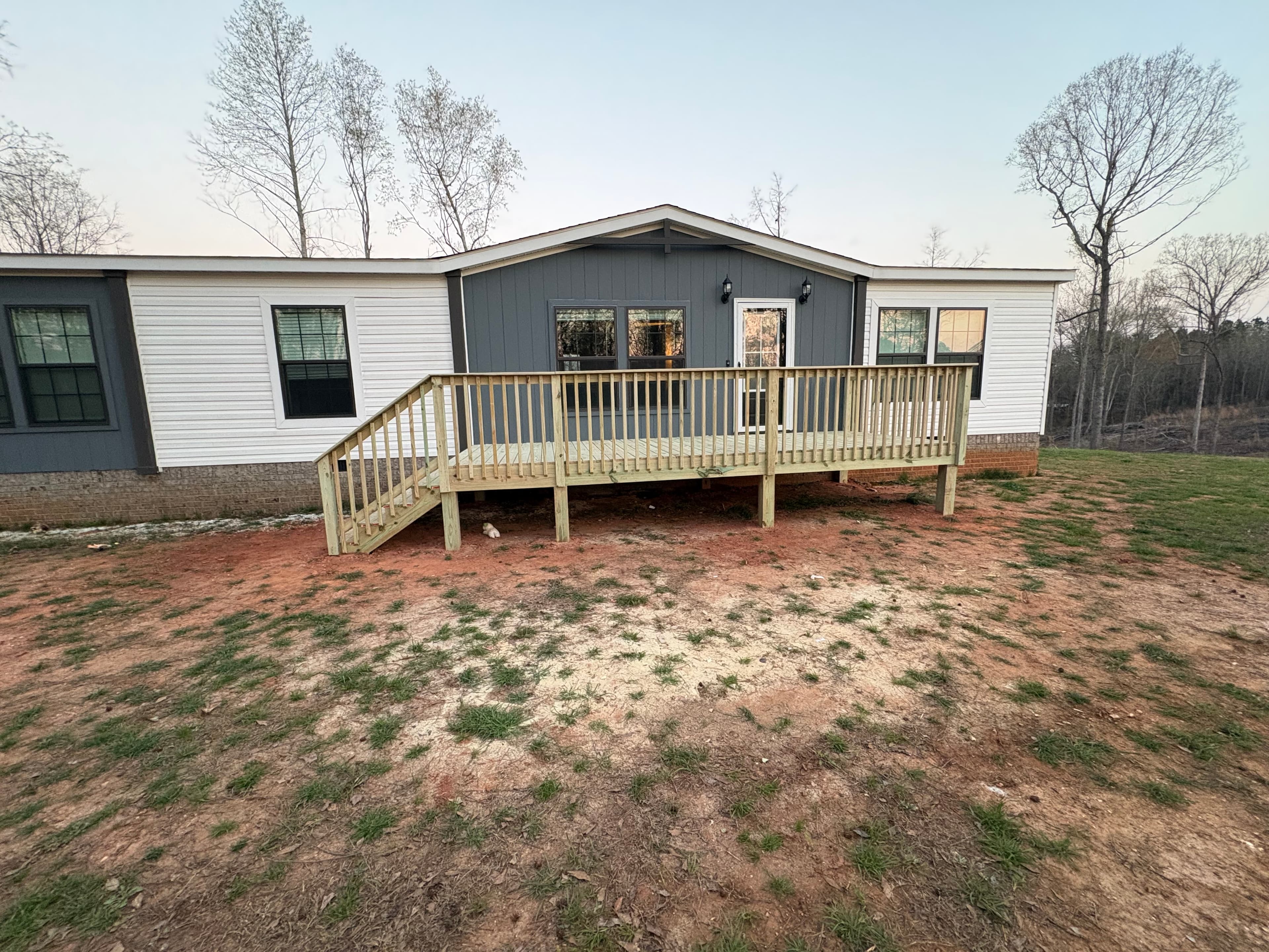 White and gray manufactured home with a wooden deck and stairs in a dirt yard.