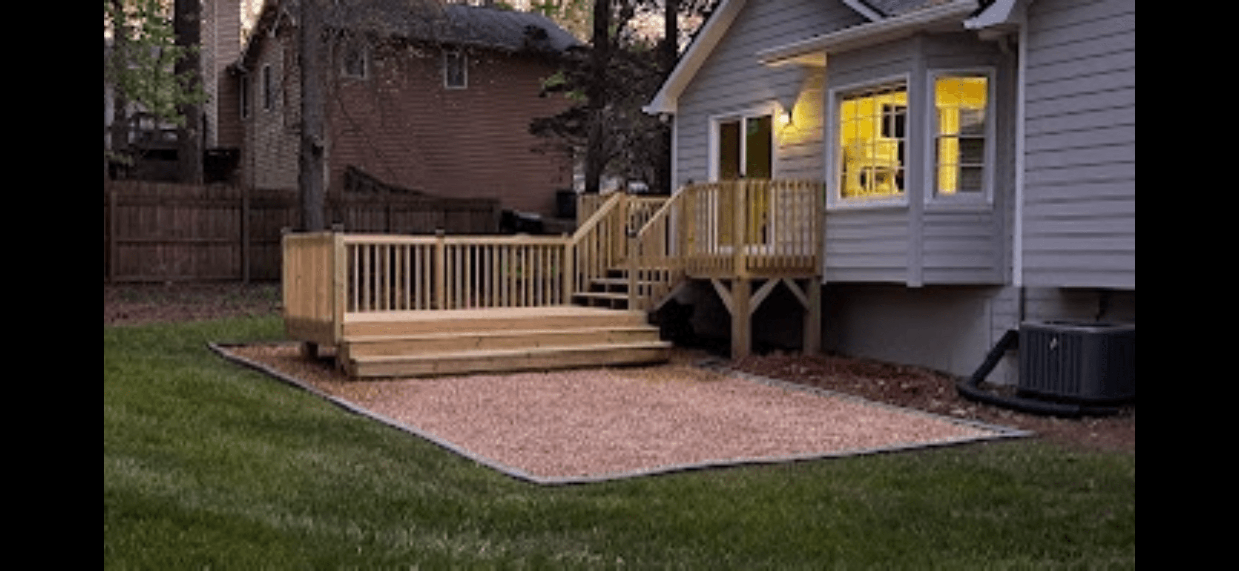 Two-tier wooden deck with stairs and railings attached to a grey house in a backyard.